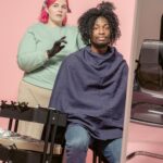 Female hairstylist with bright dyed hair doing hairstyle to African American customer wrapped in cape in studio against pink background